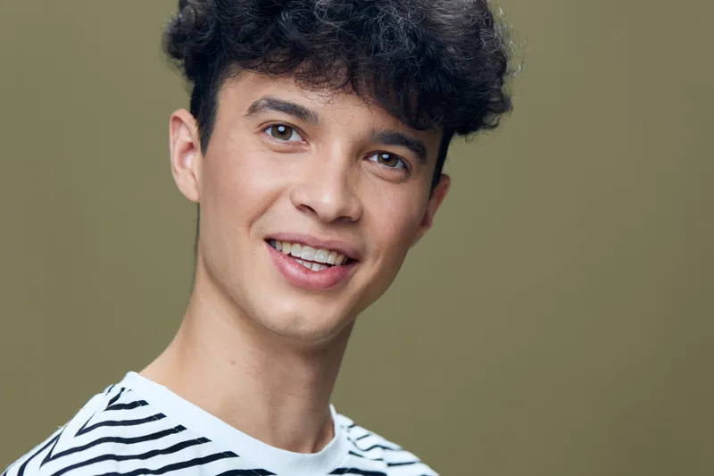 smiling man with curly hair in striped shirt looking happy and confident against neutral background, young adult portrait with natural lighting