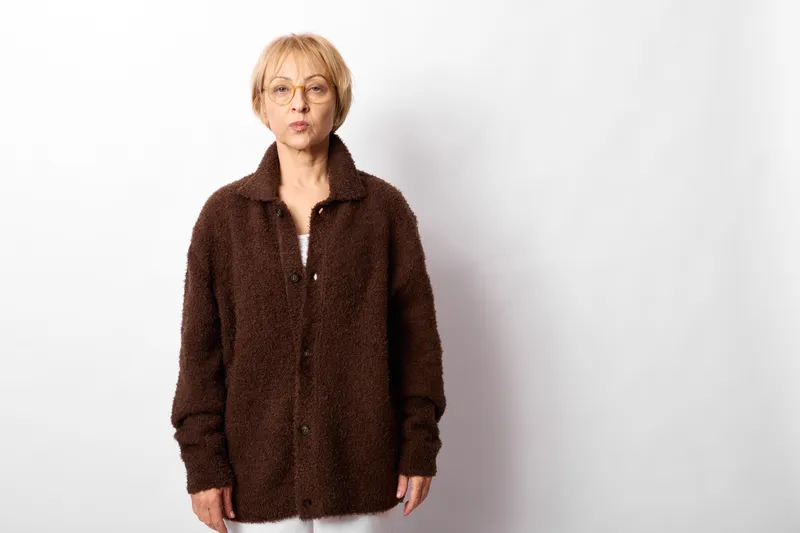 Elderly woman portrait shows serious expression, loneliness and sadness, standing indoors on a white background, looking at camera, wearing dark jacket, studio concept