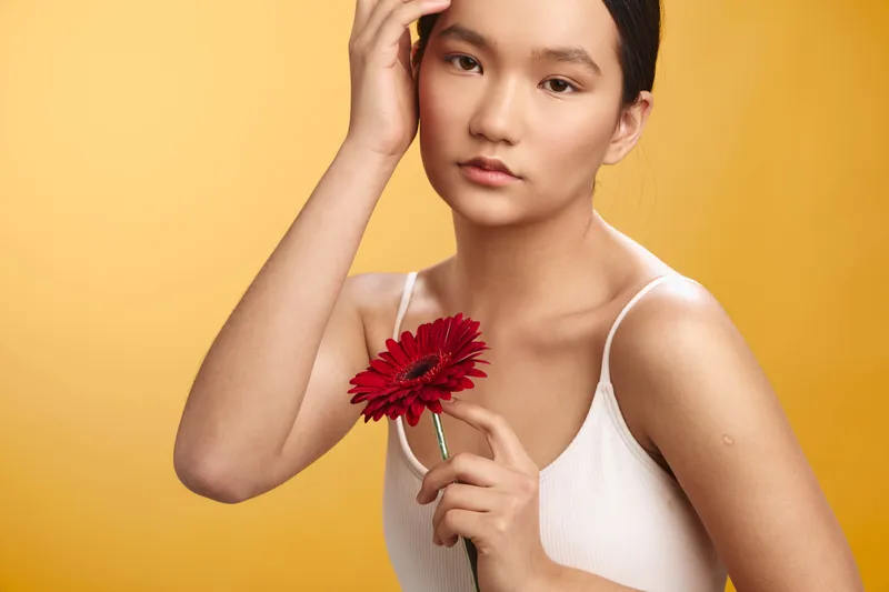 asian woman holds a red flower in a studio portrait, wearing a white top with soft lighting and a warm yellow background, creating a calm, elegant and artistic mood.