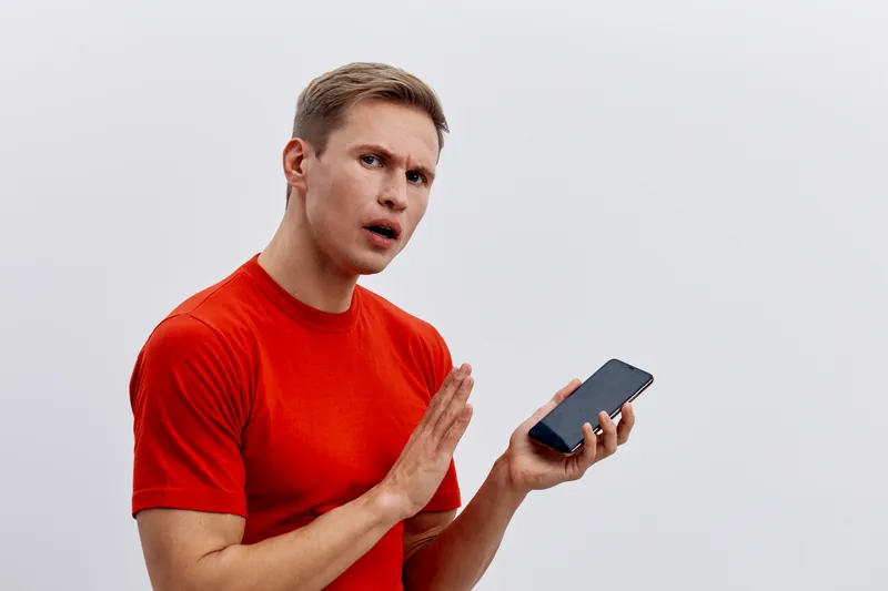 Confused young man holding smartphone with expression of surprise or disbelief, wearing bright red t-shirt, isolated on plain gray background. People emotions and technology concept.