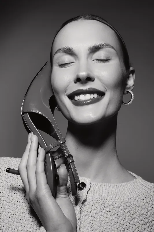 Happy Caucasian woman with fair skin smiles with closed eyes while holding a high heel shoe near her face. Concept of fashion, beauty, happiness, and footwear accessory.