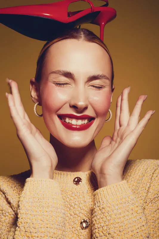 Smiling woman with red lipstick and closed eyes holds red high heel shoe on head. Fashion concept with joyful expression, yellow background, and cozy knitted sweater in studio lighting.