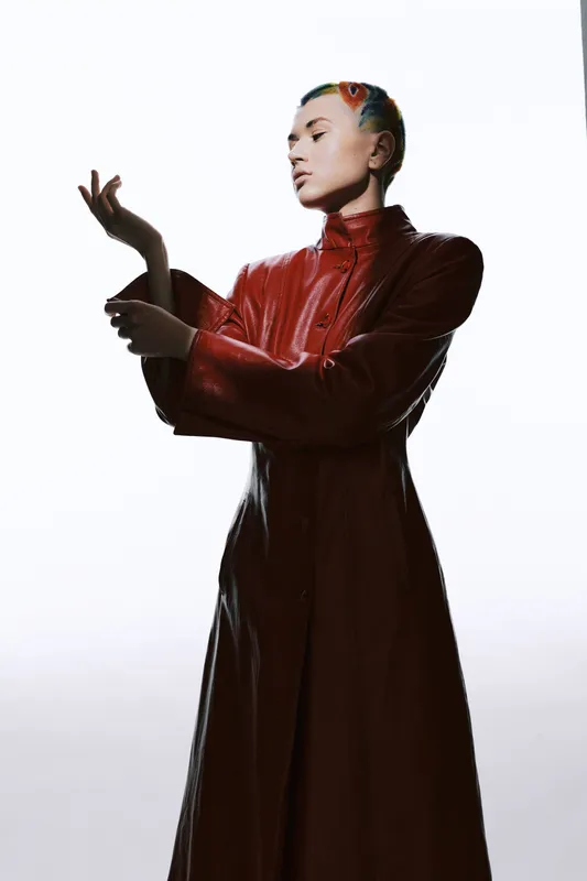 Woman with colorful hair posing confidently in a long red leather coat, showcasing modern fashion style and unique edgy look against a bright background.