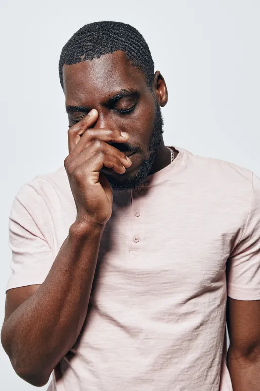 African American man with dark skin and short hair wearing light pink shirt standing with eyes closed and hand touching face in thoughtful or emotional pose on white background.