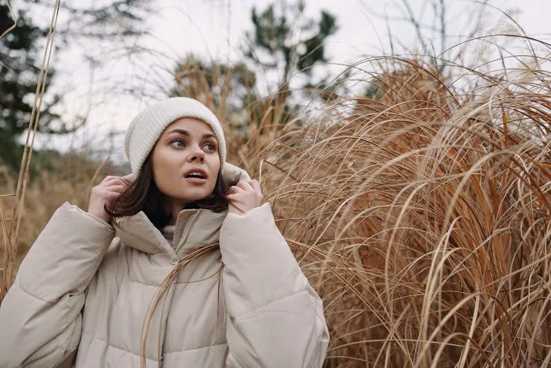 Woman in a beige puffer coat and knit beanie adjusting her hood in a chilly outdoor setting among tall dry grasses, conveying seasonal style and quiet anticipation