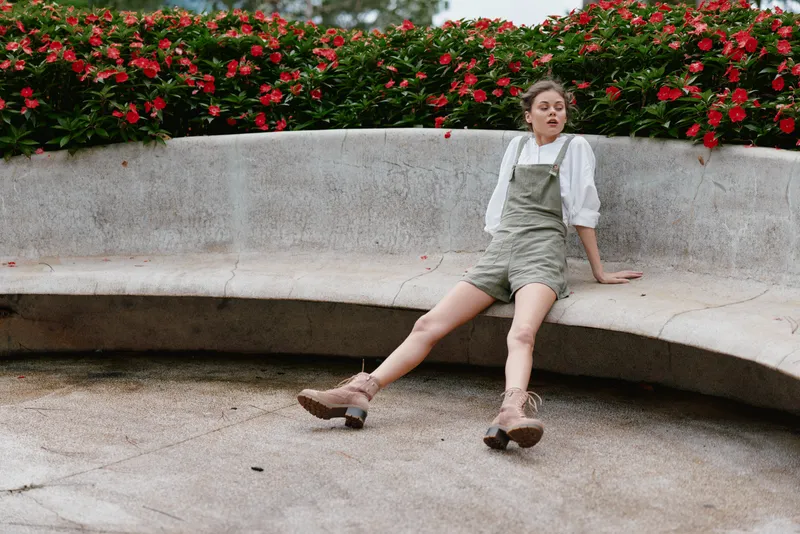 Young woman in stylish overalls and boots, relaxed pose in a serene outdoor setting with vibrant flowers and green foliage for fashion and lifestyle concepts