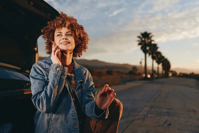 Woman with curly hair, denim jacket, standing by a car, roadside, smiling, phone, sunset glow, lifestyle concept, casual travel, outdoors