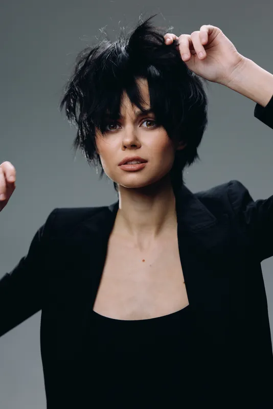 Young woman with short black hair and black blazer posing with serious expression indoors