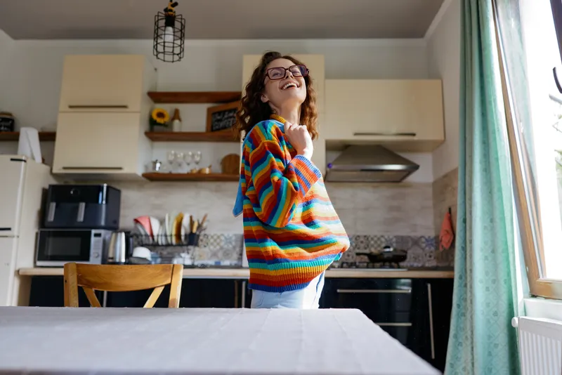 A joyful woman in a colorful striped sweater stands in a bright kitchen, smiling at the camera, embodying home lifestyle and casual comfort in a warm domestic scene.