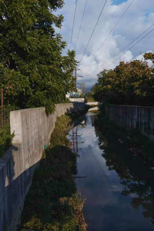 Canal, concrete wall, reflection, power lines, trees, sky — narrow urban canal with concrete walls and mirrored water, lone photographer shadow in casual outfit, autumn light and tranquil mood.