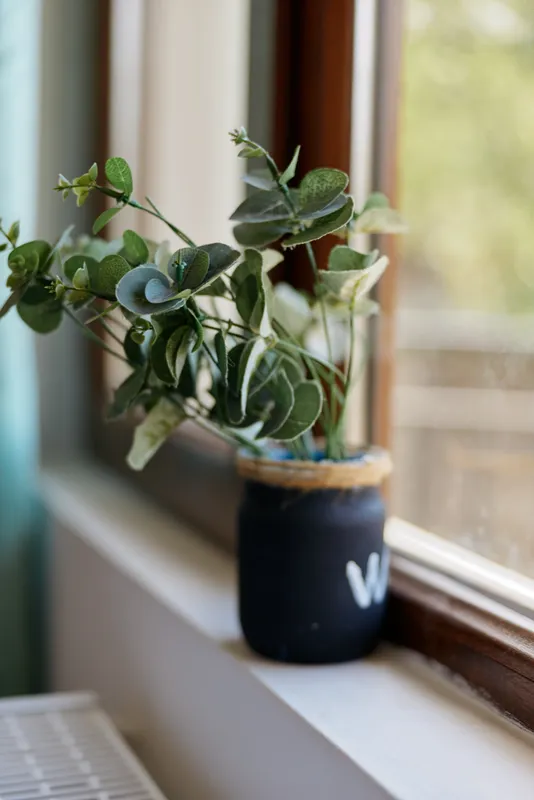 A potted plant sits on a sunlit windowsill, green leaves in a dark ceramic pot, capturing a calm, cozy home moment focused on natural greenery and simple home decor.