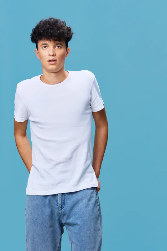 Young man in casual white t-shirt and blue jeans standing against a blue background, with a relaxed and thoughtful expression, modern fashion style.