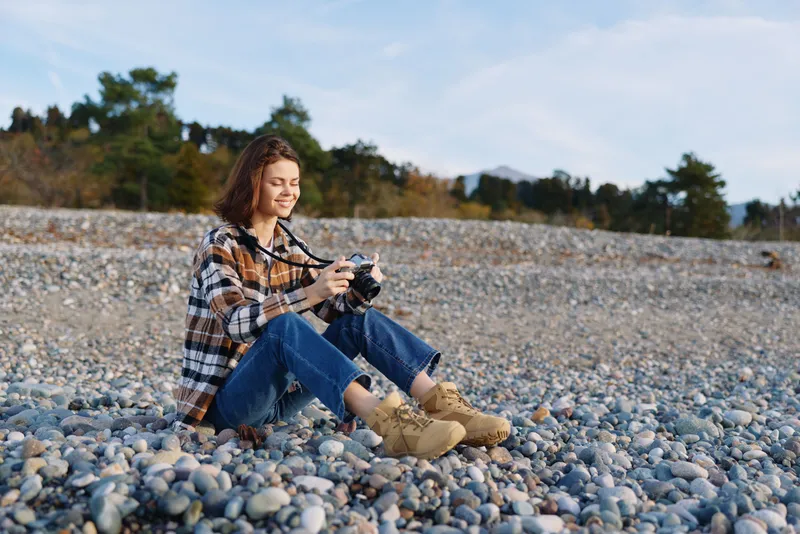 woman outdoors with camera, casual attire sitting on pebbles, beach scene showcasing nature, outdoor photography and hobby, relaxed mood and scenic daylight