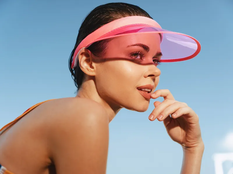 Closeup portrait of a stylish woman wearing a pink plastic visor under a bright sun, sunlit skin, relaxed smile, and confident pose against a clear blue sky.