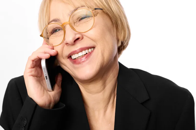 Woman smile glasses phone talking senior communication portrait studio background, closeup of a friendly mature blond woman in blazer, holding mobile phone indoors