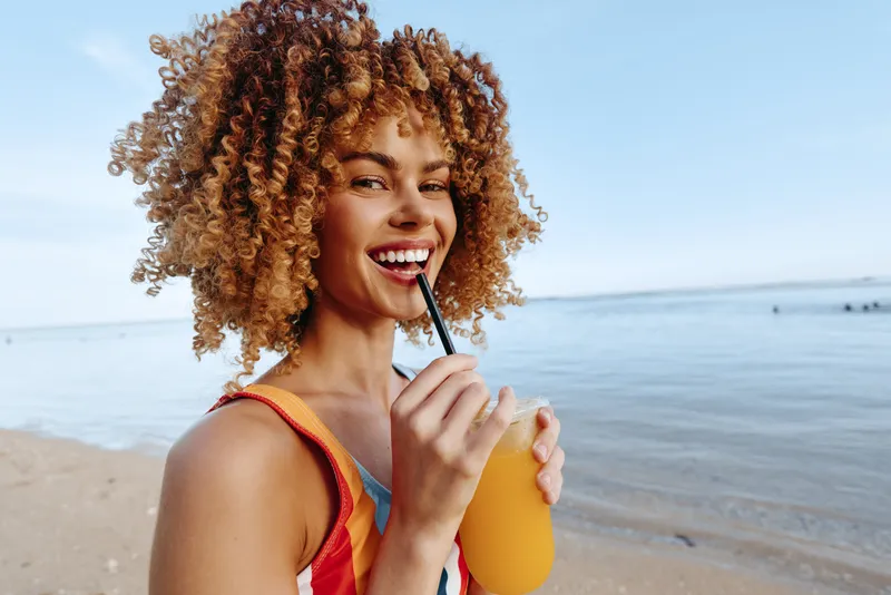 A smiling woman drinks orange juice through a straw at the sunny beach, wearing a bright tank top. A lifestyle moment capturing summer refreshment and carefree coastal vibes.