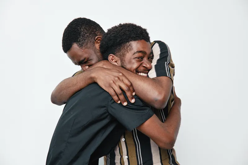Two young black men with dark skin hugging each other with smiles on white background. Friendship, happiness and connection between African ethnicity male friends in casual clothes.