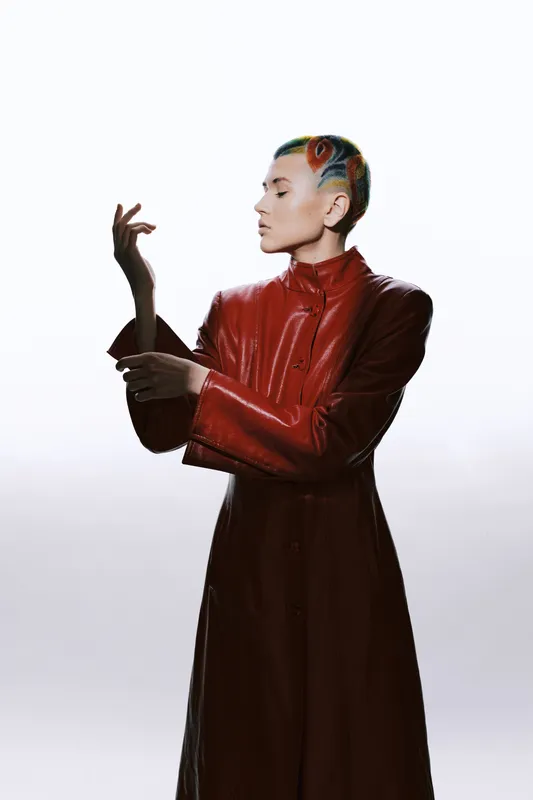 Woman with colorful short hair wearing red leather coat poses elegantly against white background, showcasing modern fashion and vibrant hairstyle.