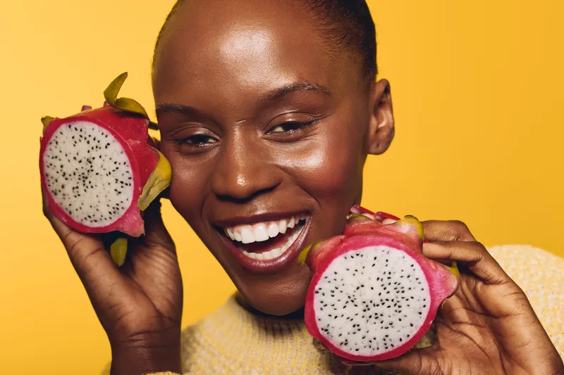 Smiling black woman with dark skin holds two halves of a dragon fruit near her face on yellow background. Concept for healthy food, tropical fruit, nutrition, and natural beauty.