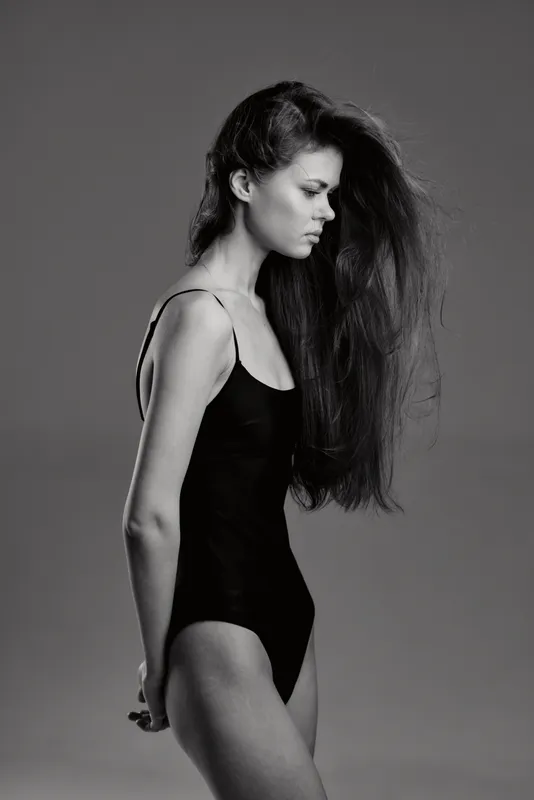 Black and white portrait of a young woman with long hair wearing a black bodysuit, looking down thoughtfully against a plain background.