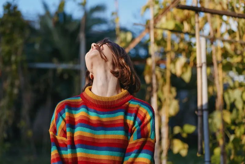 Woman in a colorful striped sweater with red hair looking up and smiling outdoors in a tropical garden, casual outfit and relaxed mood, lifestyle travel portrait in soft sunlight