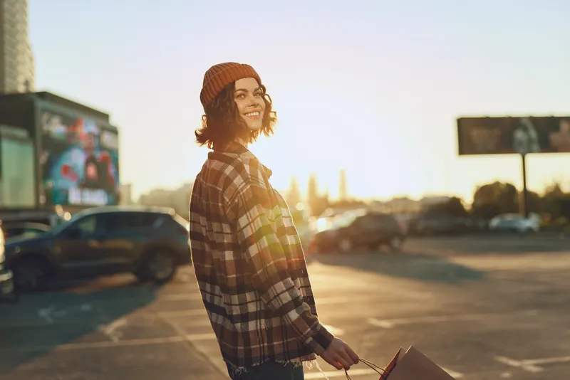 Young person with a warm smile holding shopping bag in urban street, candid lifestyle portrait in beanie and backlight, golden hour glow, authenticity and emotional storytelling vibe.