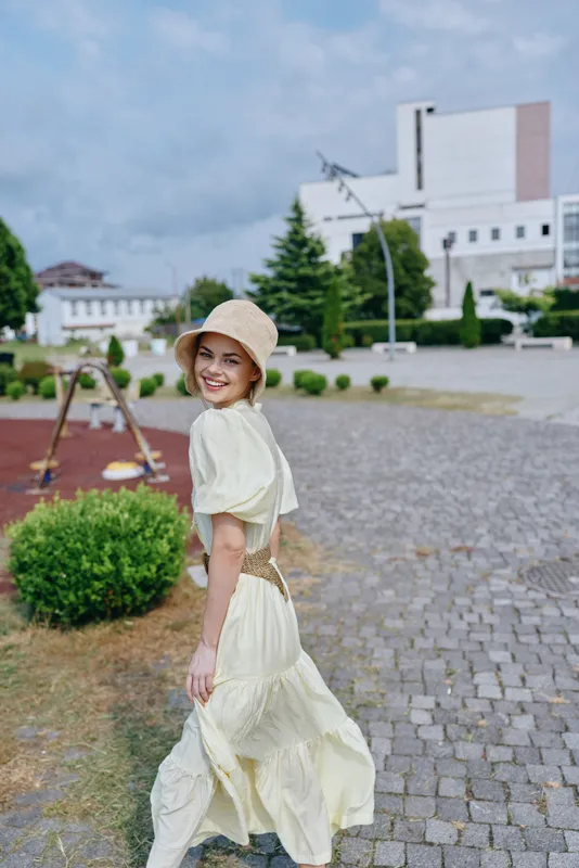 Woman in a cream dress and straw hat walks outdoors on a sunny day, smiling, along a paved path with contemporary buildings in the background