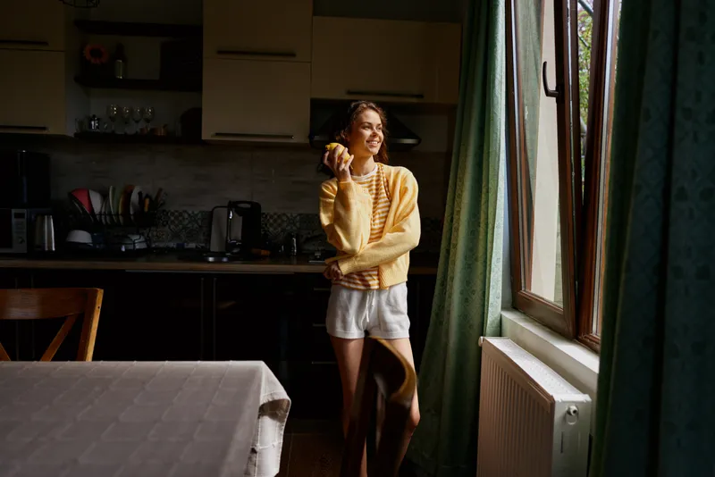 A woman in a cozy kitchen by a sunlit window, wearing a yellow sweater and shorts, smiles while holding an orange. A warm home lifestyle moment.