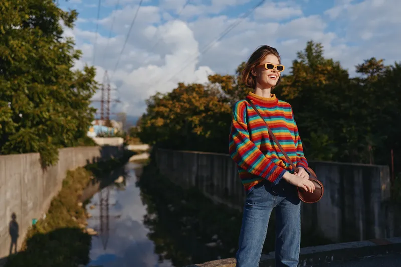 Young woman in a colorful striped sweater with sunglasses and denim jeans stands by an urban canal, casual outfit and small round bag, smiling for travel lifestyle and street fashion.
