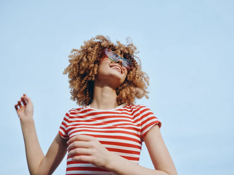 Young person with curly hair smiles under a clear blue sky, wearing a red and white striped shirt and sunglasses, radiating carefree summer energy and playful confidence.
