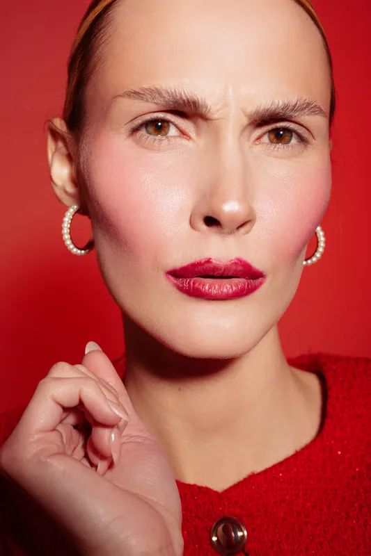 Close-up portrait of a woman with red lipstick and hoop earrings on red background. She wears a red sweater and shows a serious facial expression with furrowed brows. Fashion and beauty concept.