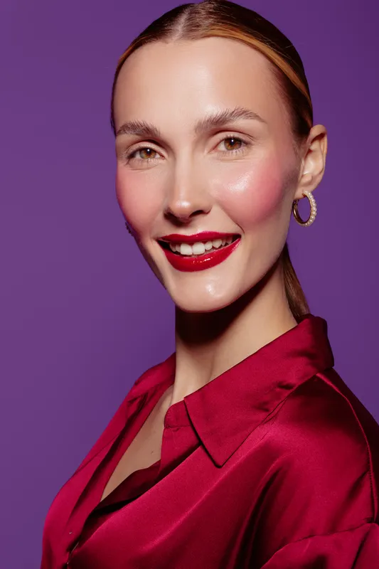Smiling woman with red lipstick and shiny skin wears red satin blouse and gold hoop earrings. Portrait on purple background. Concept of beauty, fashion, and confident female model.