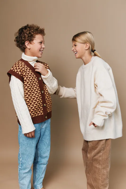 Two young friends share a warm moment in cozy knitwear, smiling and talking as they stand against a neutral backdrop, showcasing casual style and honest friendship.