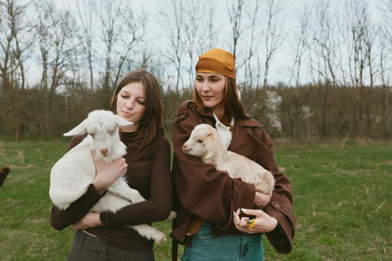 Two women outdoors, smiling softly while holding goat kid companions, casual jackets sway in a light breeze, sunlit countryside scene, natural colors, peaceful moment, friendly connection, rustic