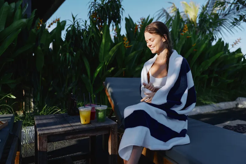 Relaxed woman sits outdoors wrapped in a striped towel, enjoying sunny weather beside drinks on a wooden table, capturing a wellness oriented lifestyle moment of self care