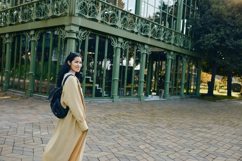 Woman with backpack and long coat walking beside glass architecture in an outdoor courtyard, showcasing fashion, travel style and urban exploration photography