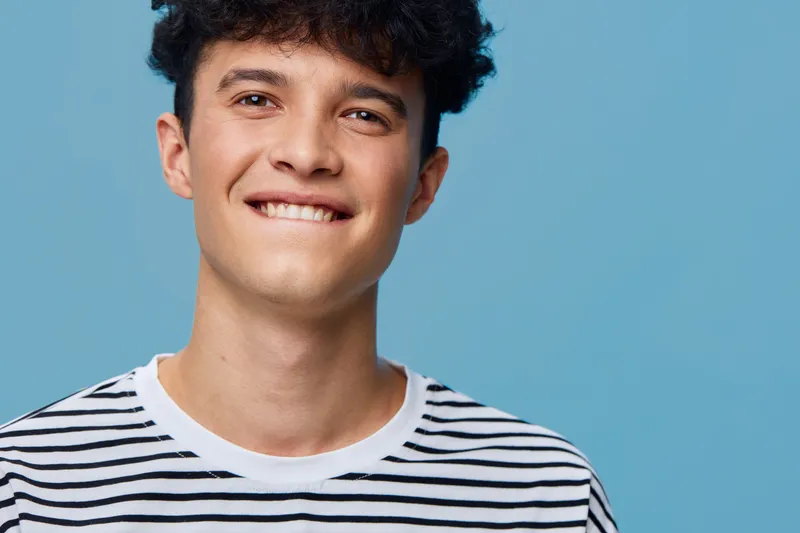 Smiling young man wearing striped shirt posing against blue background with playful expression, dark curly hair, casual style, and bright lighting.