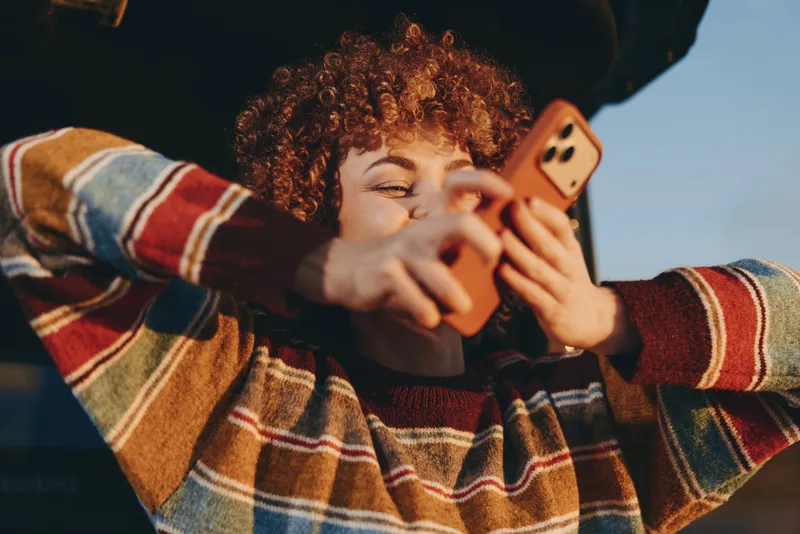 Woman lifestyle scene, curly hair woman relaxing on couch, plaid sweater, smiling while holding smartphone, taking selfie, warm evening light, casual home interior