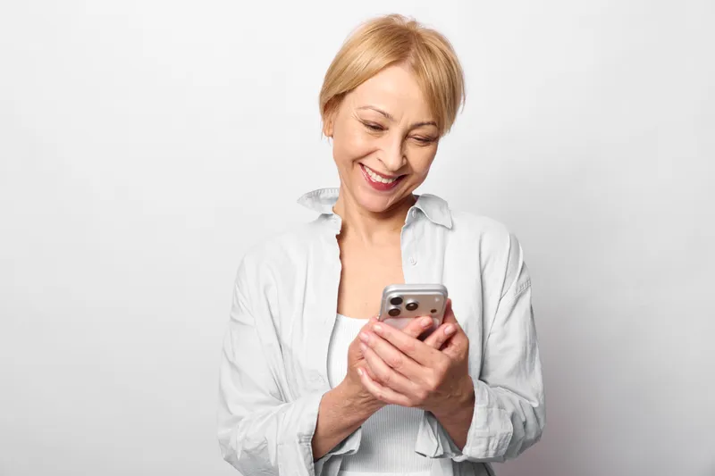 Smiling mature woman with smartphone, looking at screen in studio portrait, white background, relaxed posture, casual shirt, warm expression, mobile communication technology