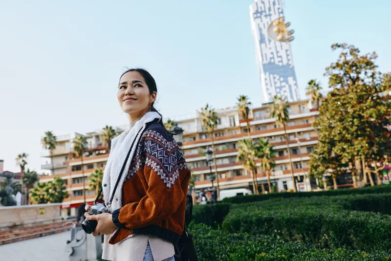 Woman wearing scarf and knit sweater holding camera in an urban park with palm trees and modern buildings, capturing street style and travel mood during daytime.