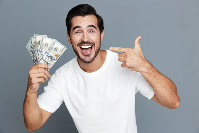 Happy cheerful young man holding cash in hand, smiling broadly while pointing at money. Isolated on solid gray background. Joy of finance concept.