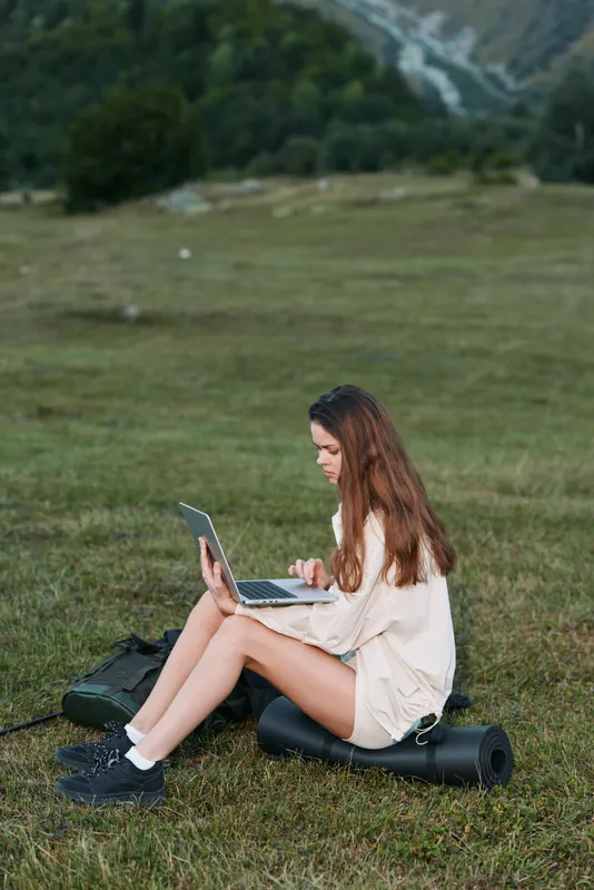 A young woman sits on a grassy field, typing on a laptop with a calm, focused expression. The open landscape creates a serene, productive outdoor work scene.