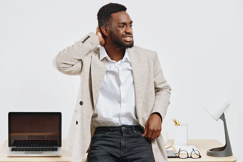 Professional young black man in casual jacket and white shirt stands at desk with laptop and stationery in modern office interior, looking away while adjusting hair, cheerful mood, business casual sty