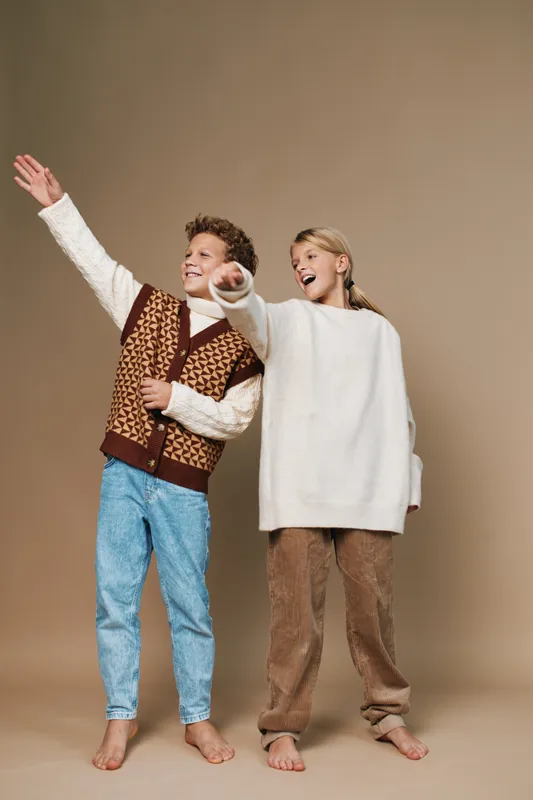 Two cheerful children pose with outstretched arms, smiling as if inviting adventure, dressed in warm knitwear and cozy sweaters against a neutral studio backdrop.