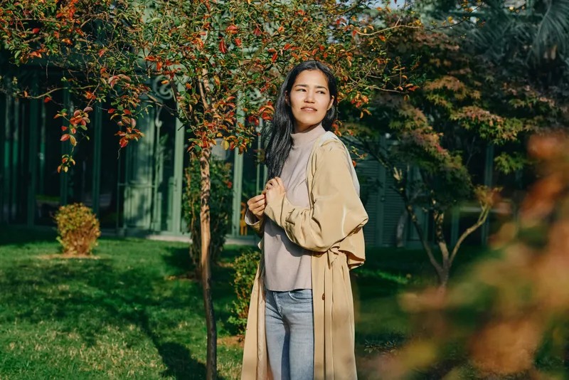 Woman in beige coat stands near blooming trees in a sunny park, casual outdoor fashion, serene smile, soft daylight, nature backdrop, relaxed stroll, spring ambience, candid lifestyle scene