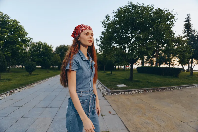 lifestyle woman in modern boho-western denim street style walks outdoors with confident expression, red bandana, and natural background in film color tones