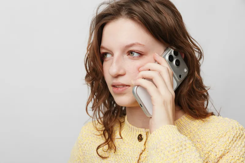 Young woman with curly hair wearing yellow sweater talks on smartphone, looking away. Concept of communication, technology, casual lifestyle, and thoughtful expression.