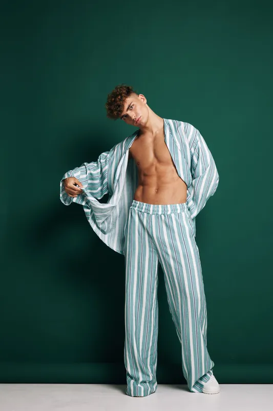 Young man wearing striped pajamas standing against dark background with open shirt revealing toned abs. Fashion portrait portrait of trendy casual male style and confident posture