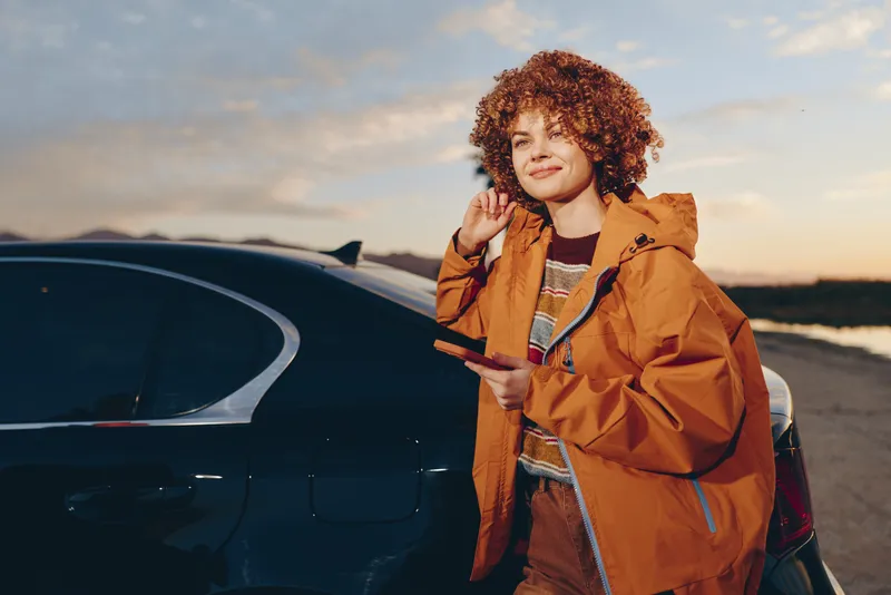 Woman lifestyle outdoors car jacket sunset curly hair road travel, orange coat woman stands near car on roadside, relaxed smile touching hair, warm dusk light, scenic horizon