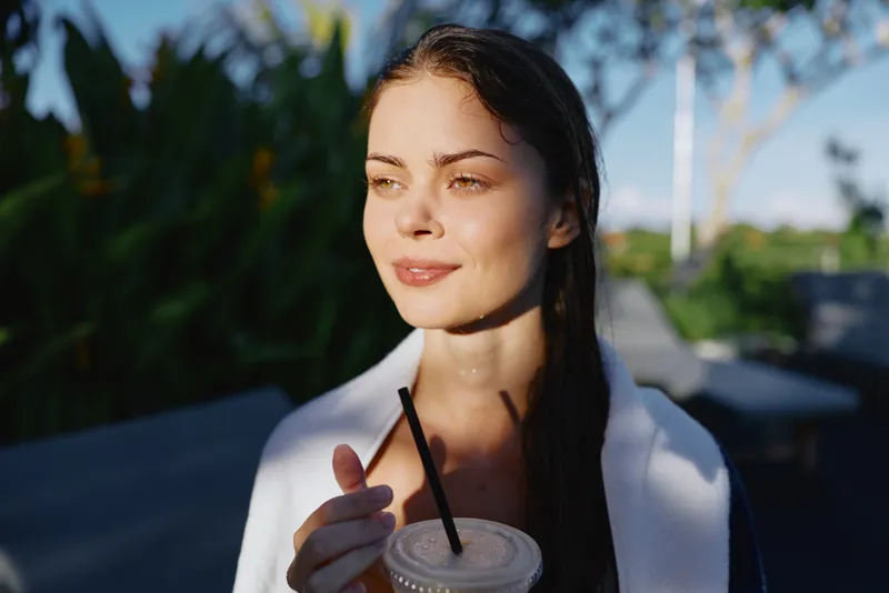 Young woman with long hair holding a cold drink with a straw outdoors. Natural sunlight highlights her relaxed expression and casual lifestyle concept.